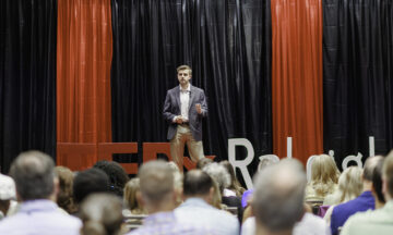 A well-dressed young man in a grey blazer and khakis giving a presentation on a stage. Behind him is a large "TEDxRaleigh" sign against a backdrop of red and black curtains, and an audience is visible in the foreground.