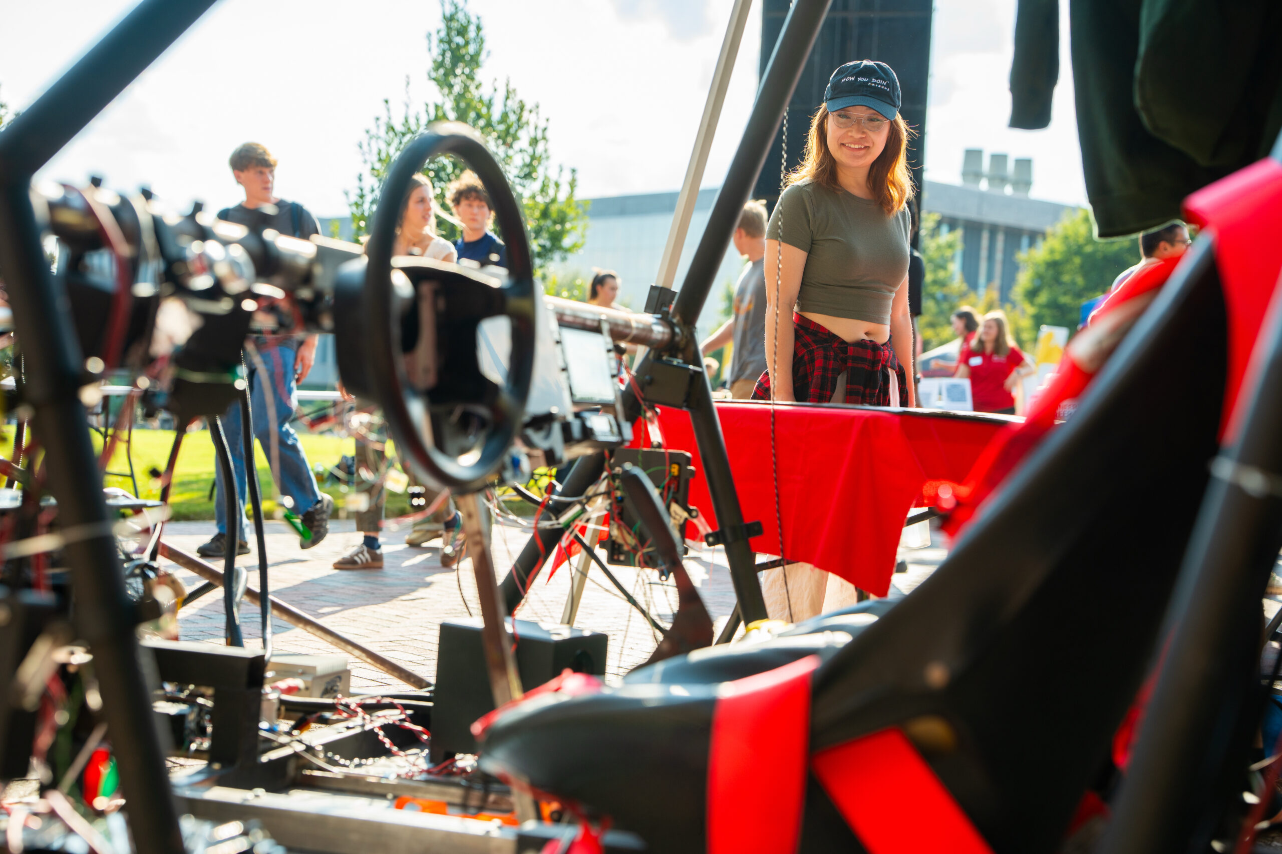 A photo of a female student. She is outside looking at a Baja motorsports car in the foreground.