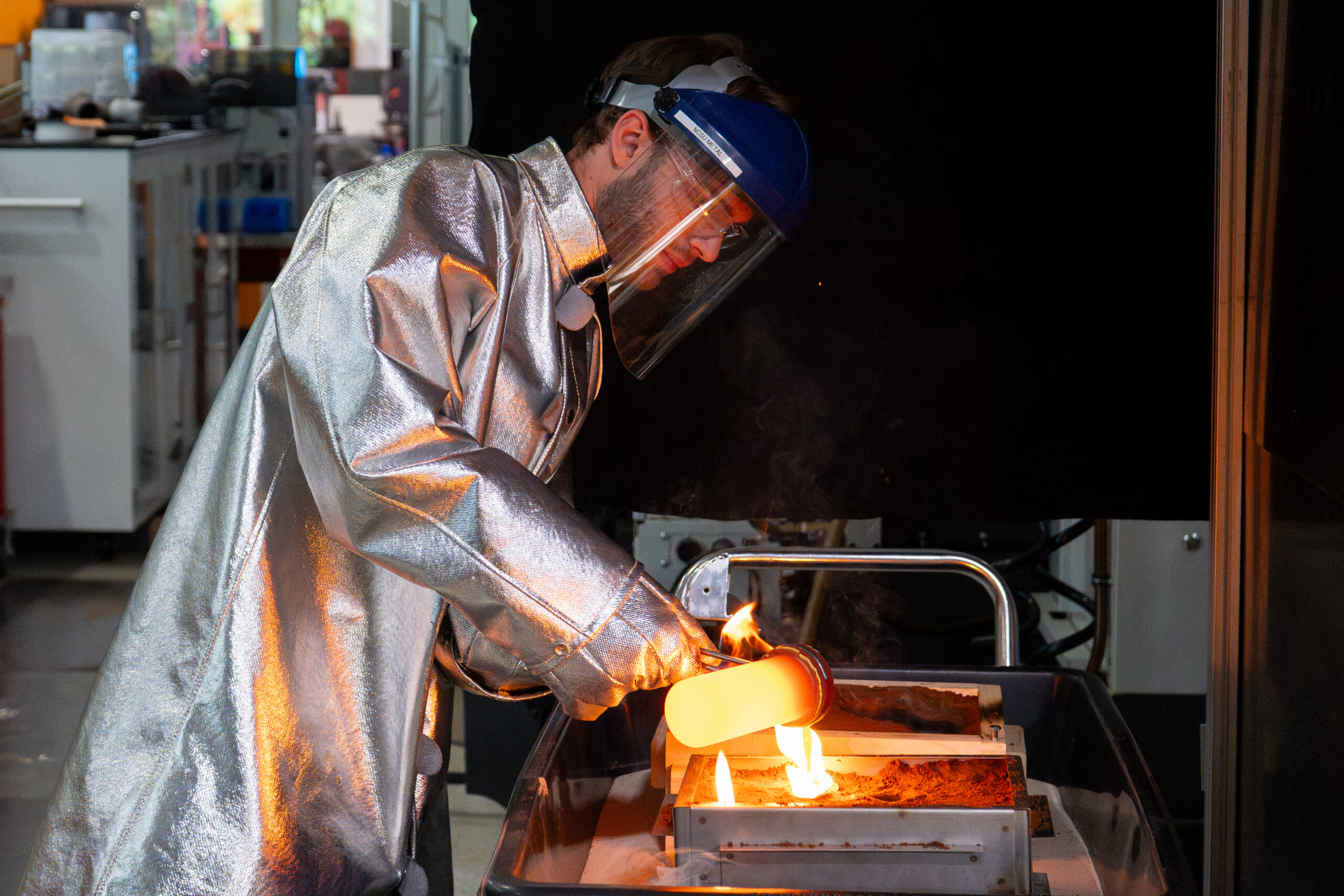 Image shows a man wearing silver safety gear and a face shield pouring melted metal.