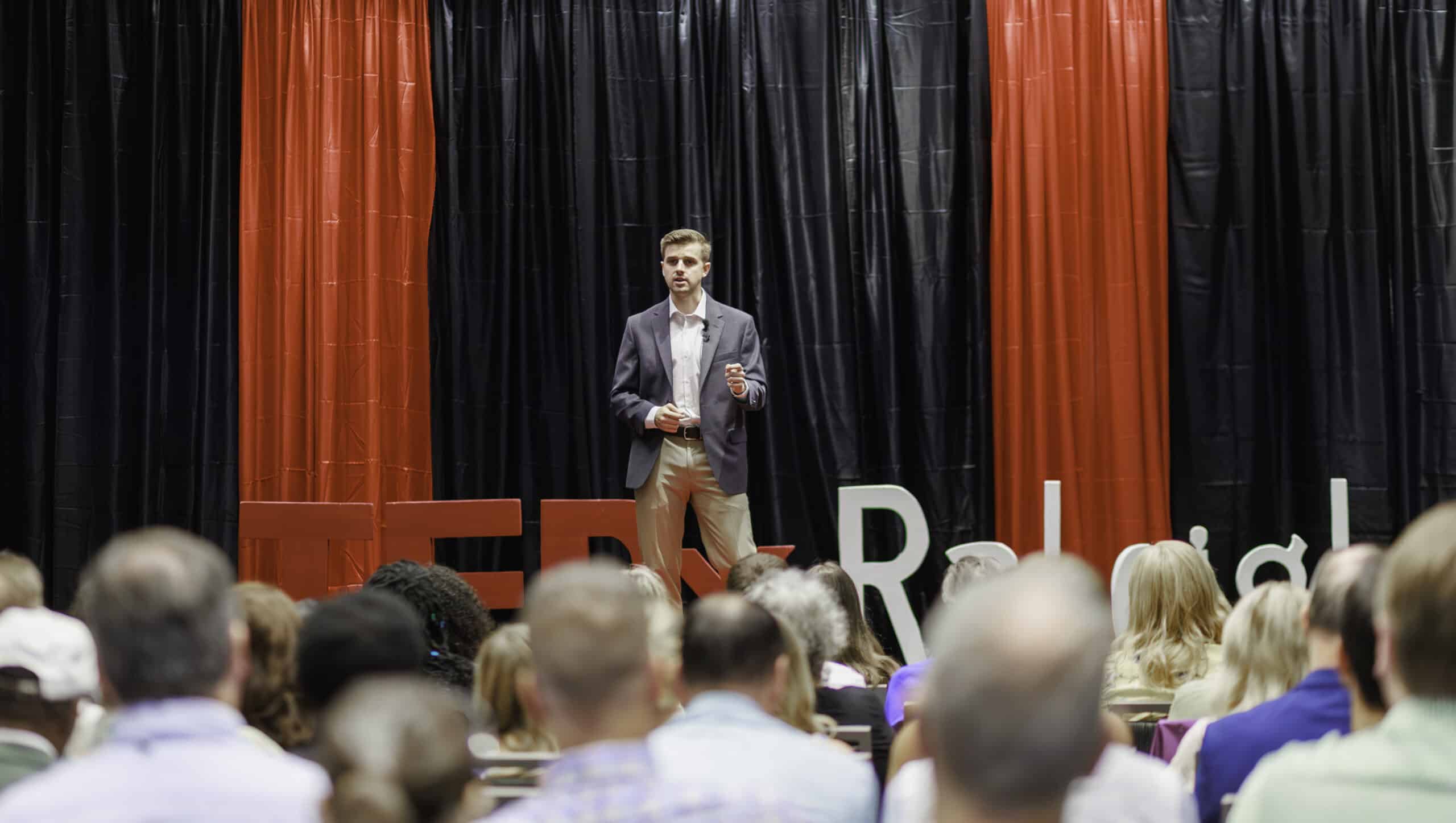 A well-dressed young man in a grey blazer and khakis giving a presentation on a stage. Behind him is a large "TEDxRaleigh" sign against a backdrop of red and black curtains, and an audience is visible in the foreground.