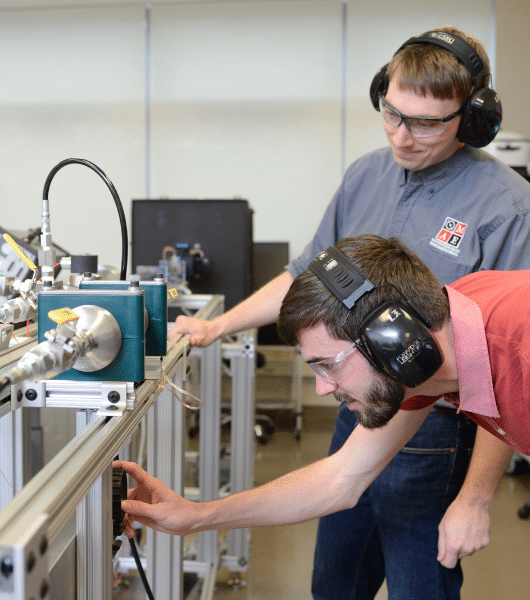 Image shows two young men wearing headphones and eye protective gear working on a piece of metal equipment.