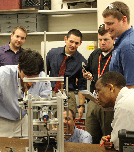 Image shows a group of professionally dressed men working on a metal machine in a classroom setting.