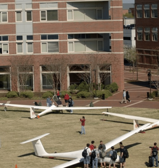 Image shows a brick building on NC State's campus with small airplane models and students on the lawn.