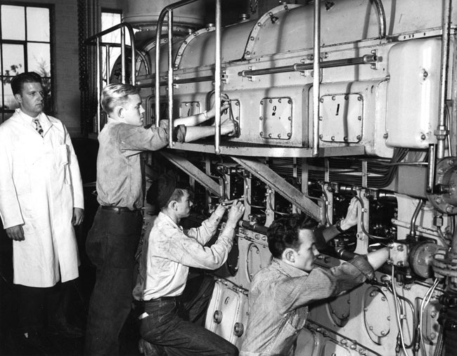 A black and white image of one man in a lab coat and three men working on a machine.